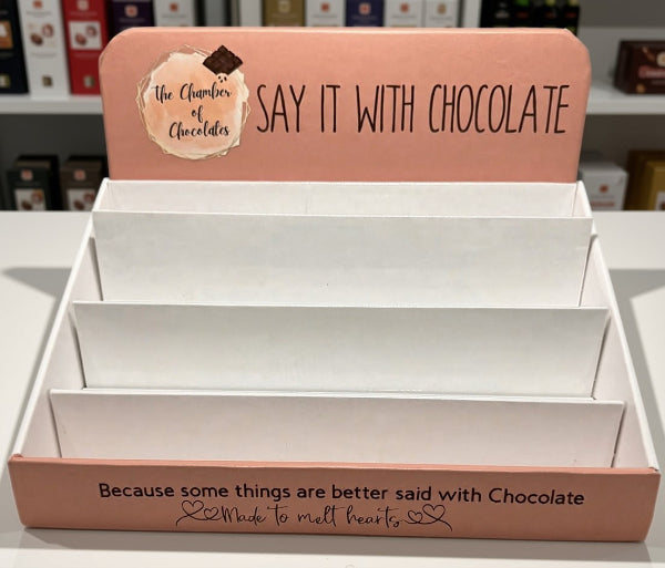 A pink cardboard display stand with white interior shelves, printed with the text 'SAY IT WITH CHOCOLATE' and 'Because some things are better said with Chocolate Made to melt hearts.' The top panel also shows a logo reading 'the Chamber of Chocolates.